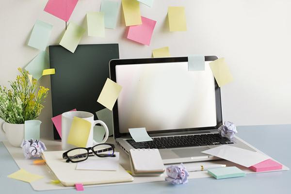 A laptop sits on a desk alongside glasses, note pads, and a mug; multiple sticky notes in assorted colors are stuck to the wall adjacent to the desk.